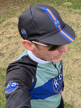 Load image into Gallery viewer, A man wearing a black cycling cap and matching cycling jersey with blue and gold stripes and A-D logos, photographed outdoors on grass.
