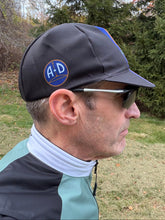 Load image into Gallery viewer, A man in profile wearing a black cycling cap with a blue and white circular 'A+D' logo patch, sunglasses, and a teal and black athletic jersey, set against an outdoor background of grass and trees.

